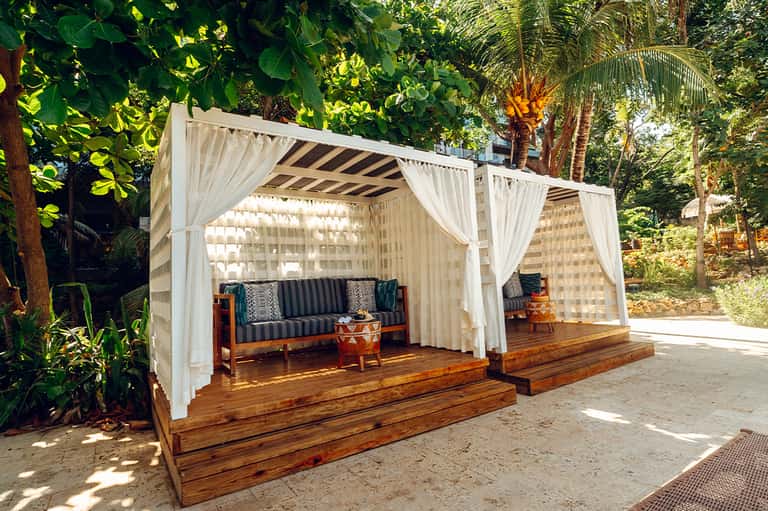 Tropical resort cabana with white curtains, wooden deck, and dark cushioned seating surrounded by lush greenery