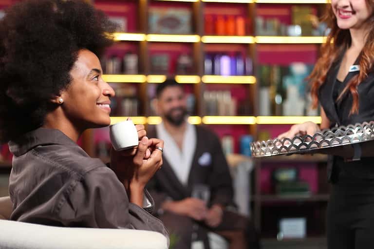 Man holding coffee cup chatting with female bartender at illuminated bar with shelves of bottles