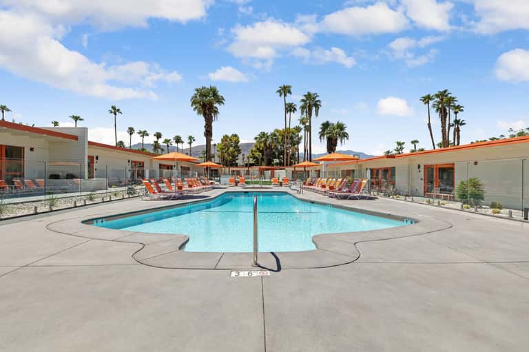 Resort pool with crystal-blue water, orange lounge chairs, palm trees, and mid-century modern buildings