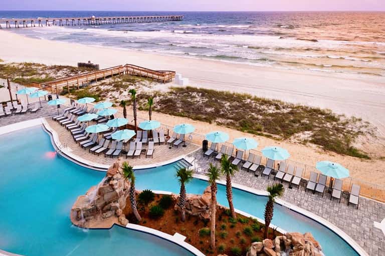 Beachfront resort with curved pool, turquoise umbrellas, lounge chairs, and pier extending into ocean