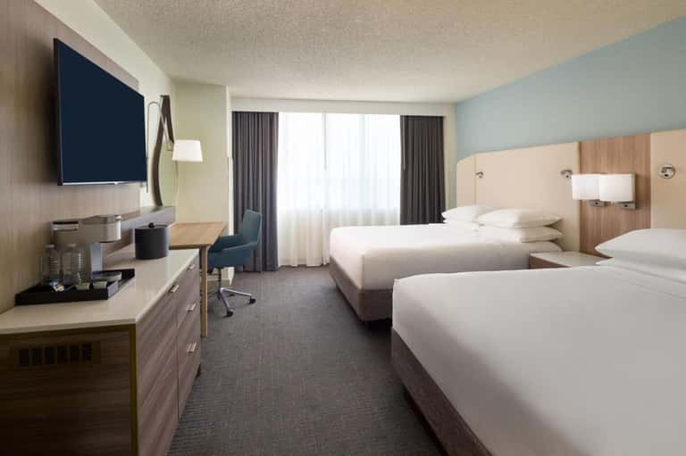 Modern hotel room with two queen beds, TV, desk, and large windows with curtains