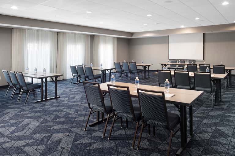 Spacious meeting room with multiple tables, black chairs, projection screen, and natural light