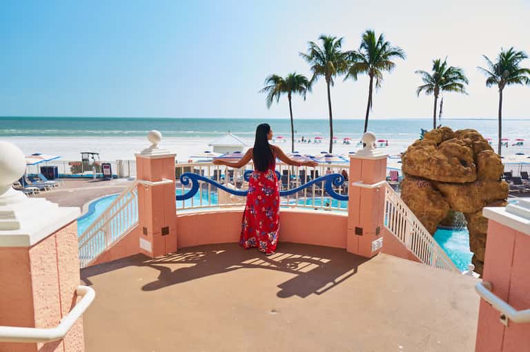 Woman in red floral dress stands on terrace railing overlooking beach with palm trees and ocean