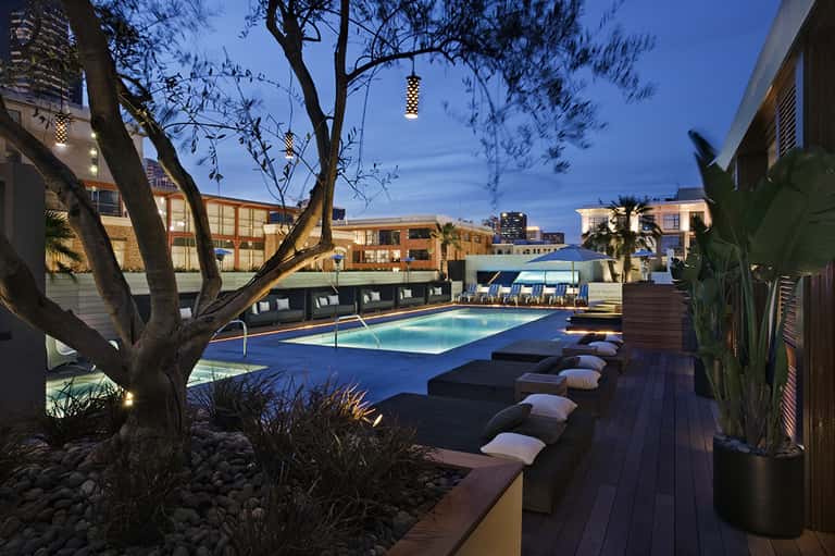 Modern rooftop pool at dusk with lounge beds, city skyline, and dramatic tree lighting