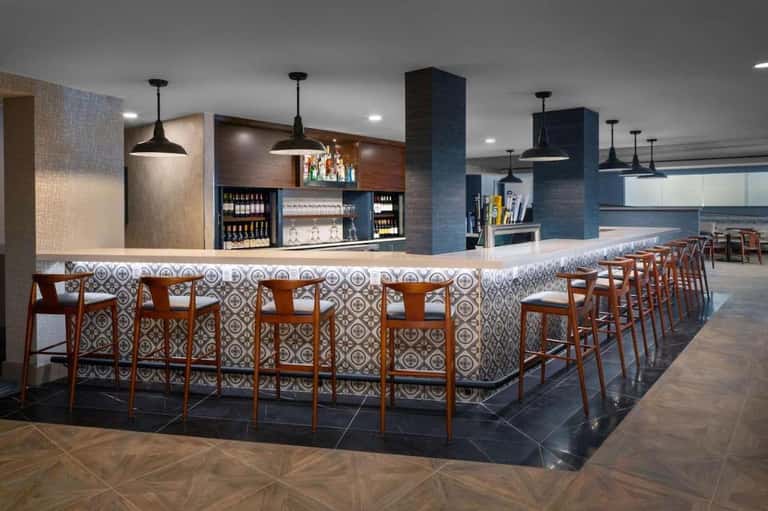 Contemporary hotel bar with patterned tile accents, wooden bar stools, pendant lighting, and shelved beverages