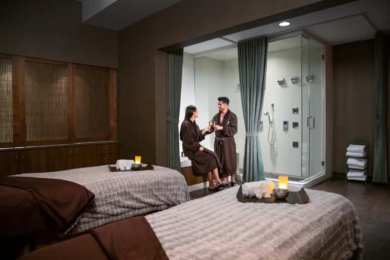Spa treatment room with two massage beds, large shower enclosure, and attendants in robes