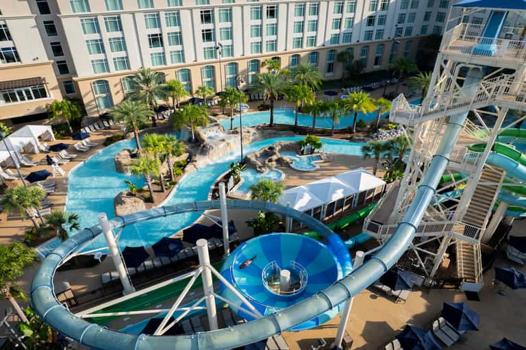 Aerial view of resort pool complex with water slides, palm trees, and surrounding hotel buildings