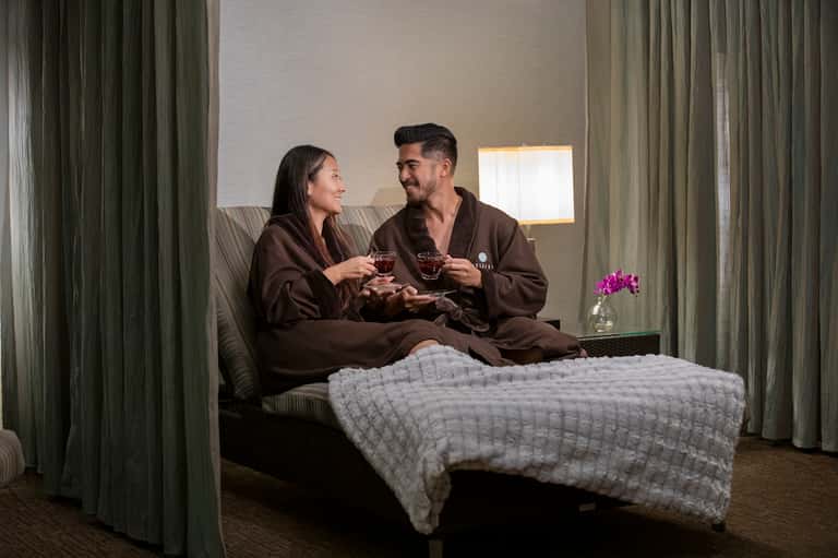 Couple in brown robes toasting with drinks while sitting on bed in modern hotel room