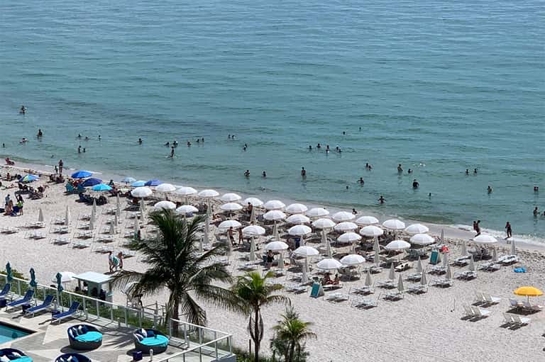 Beachfront with rows of white umbrellas, lounge chairs, swimmers in turquoise ocean, and palm trees
