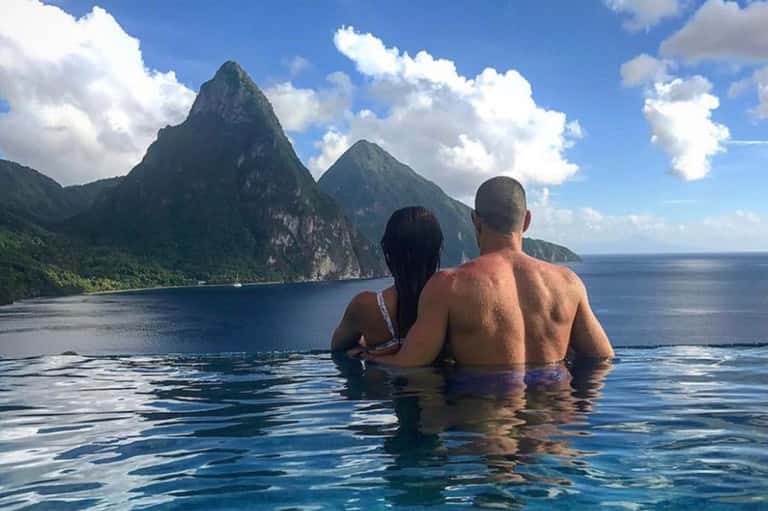 Couple swimming in crystal clear water with dramatic green mountain peaks in background under blue sky