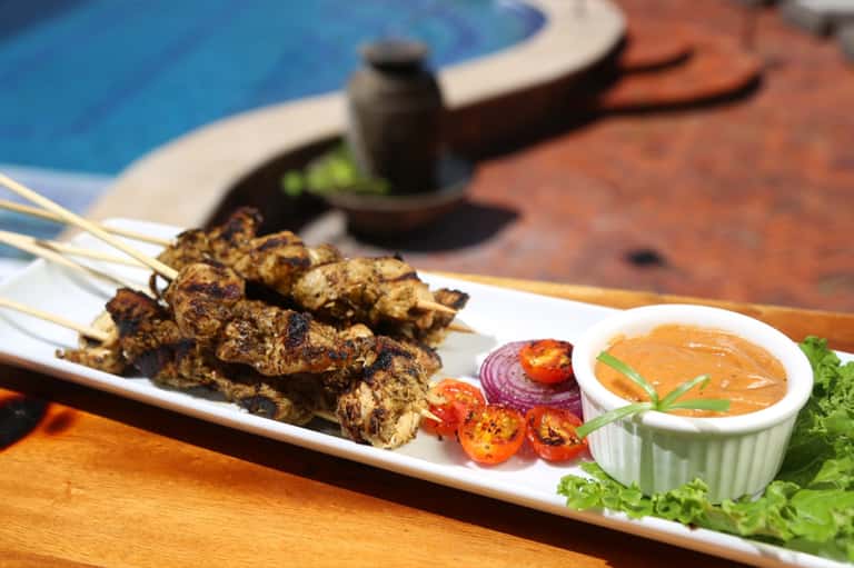 Grilled meat skewers with sauce and vegetables on white plate by poolside