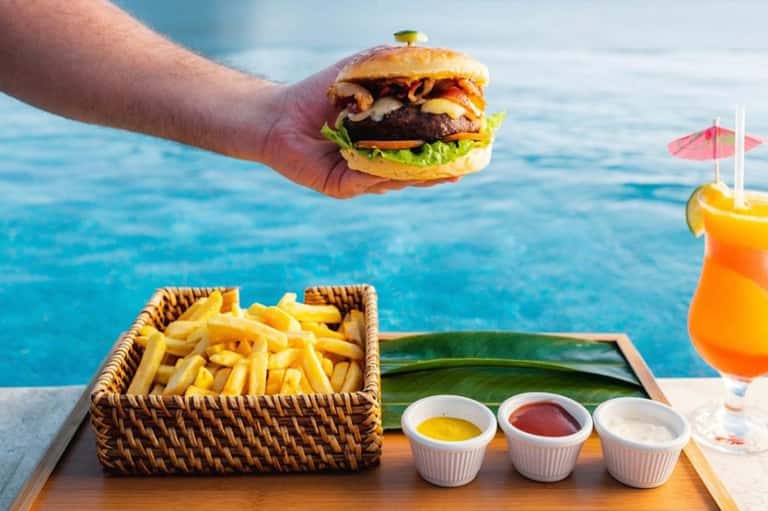 Hand holding gourmet burger with fries and tropical cocktail overlooking ocean