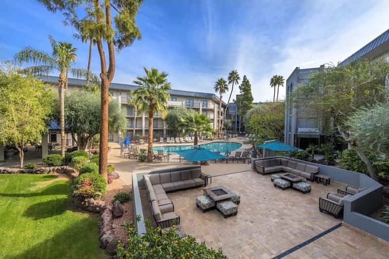 Resort courtyard with curved seating areas, fire pit, pool, palm trees, and modern buildings