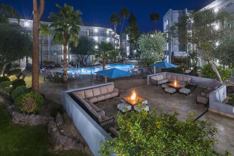 Resort courtyard at night with illuminated pool, fire pits with seating, palm trees, and modern white buildings