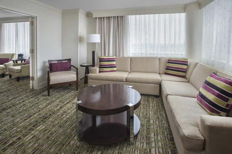 Spacious hotel suite with beige sectional sofa, round coffee table, and city views