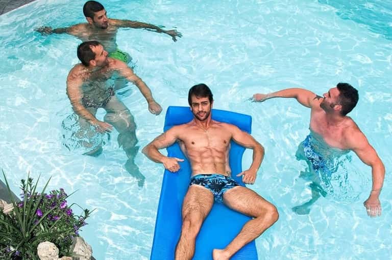Four young men enjoying a swimming pool, one relaxing on blue float while others swim nearby in crystal clear water