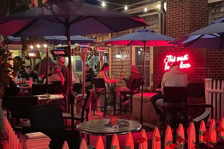 Evening rooftop bar scene with burgundy umbrellas, neon signage, brick walls, and guests at tables under warm lighting