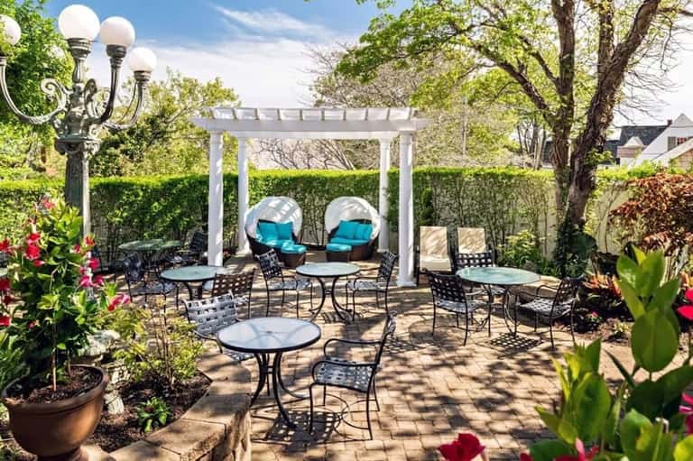Courtyard with white pergola, blue cushioned lounge chairs, metal patio tables, flowering plants, and lamp post