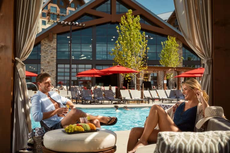 Couple relaxing on lounge chairs by pool under wooden pergola with red umbrellas and resort building