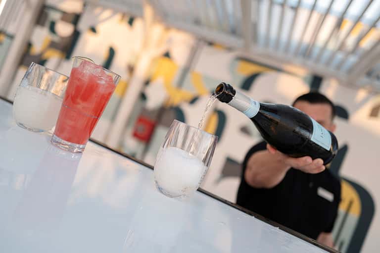 Champagne bottle pouring into glass with red and clear beverages on outdoor patio