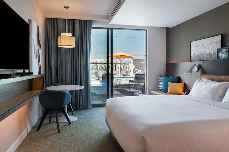 Modern hotel bedroom with king bed, desk, flat-screen TV, and sliding glass door to ocean-view balcony