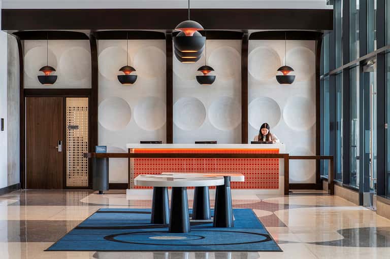 Modern hotel lobby reception desk with orange accent wall, decorative pendant lights, and geometric backdrop