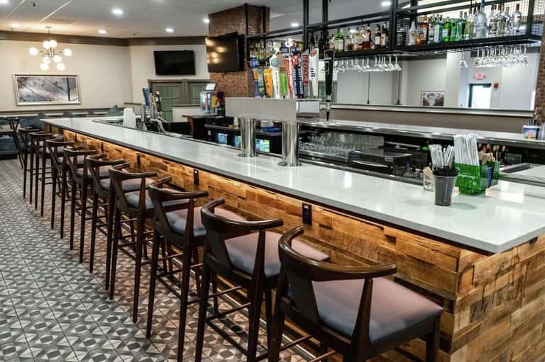Hotel bar lounge with wooden counter, bar stools, shelving with bottles, and TV on wall