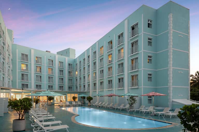 Turquoise hotel exterior at dusk with pool, loungers, umbrellas, and illuminated ground-floor spaces