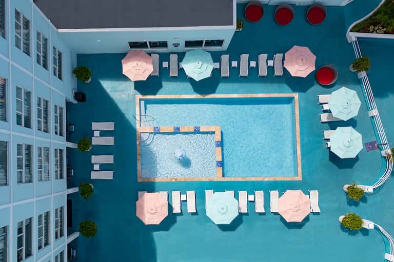 Aerial view of turquoise pool deck with pink and white umbrellas, lounge chairs, and surrounding lawns