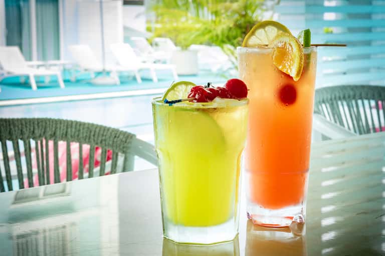 Two colorful cocktails on poolside table with lime and cherry garnishes, pool chairs visible in background