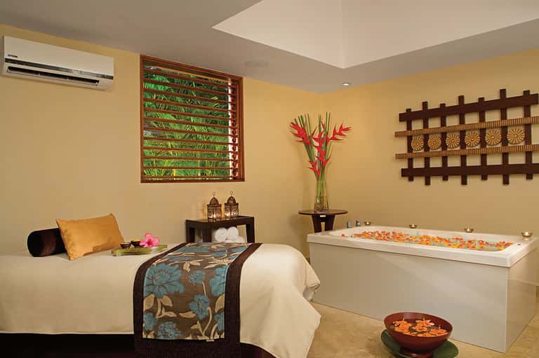 Luxurious spa room with soaking tub, massage bed, decorative flowers, and tropical-themed décor in warm gold tones