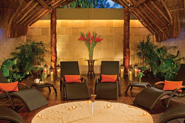 Spa relaxation room with thatched ceiling, lounge chairs, tropical plants, and candlelit ambiance