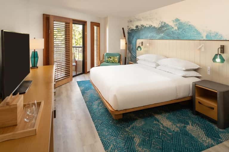 Coastal hotel bedroom with king bed, turquoise artwork, wood shutters, and balcony access