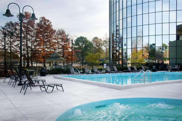 Modern hotel pool with hot tub, lounge seating, and glass building with autumn foliage backdrop