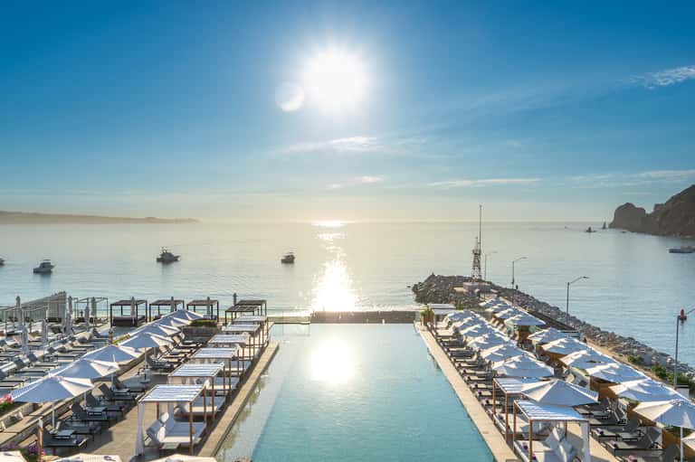 Oceanfront infinity pool with lounge chairs and blue umbrellas overlooking boats and coastal cliffs