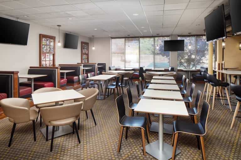 Contemporary hotel dining space with white tables, wood-frame chairs, and flat-screen TVs on walls
