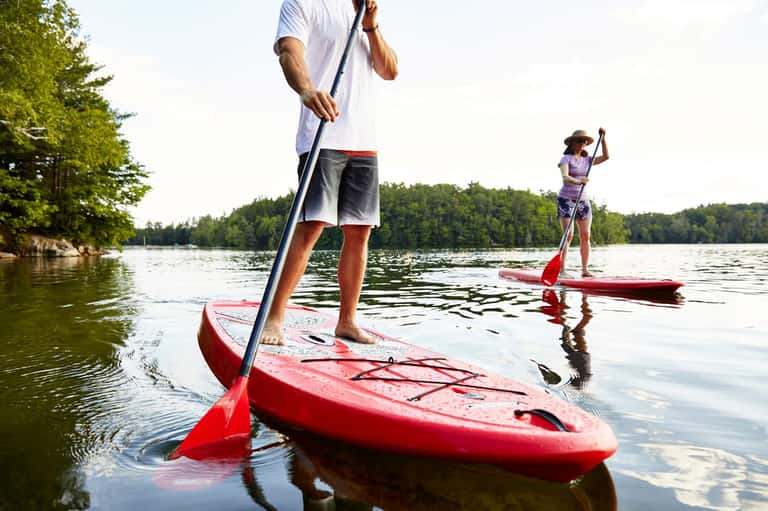 Two people paddleboarding on red kayaks in a calm lake surrounded by forested shoreline