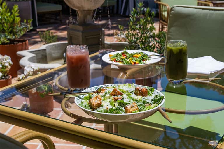 Outdoor dining table with salads, beverages, and appetizers overlooking palm-lined grounds
