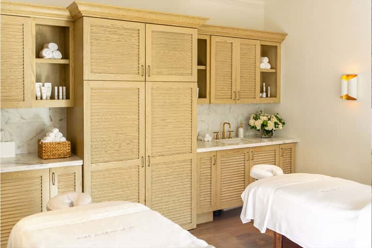 Spa treatment room with wooden cabinetry, marble countertops, two massage tables, and fresh flowers