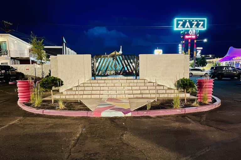 Neon ZAZZ sign illuminated at night above a pink and white geometric patterned plaza with landscaping