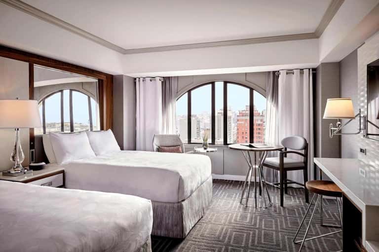 Modern hotel bedroom with two beds, city views through arched windows, and contemporary furnishings