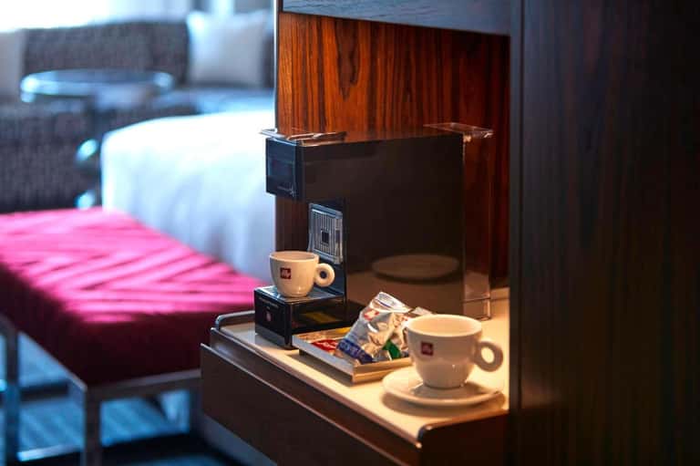Hotel room detail showing coffee maker with Illy cups on wooden desk and pink bedding in background