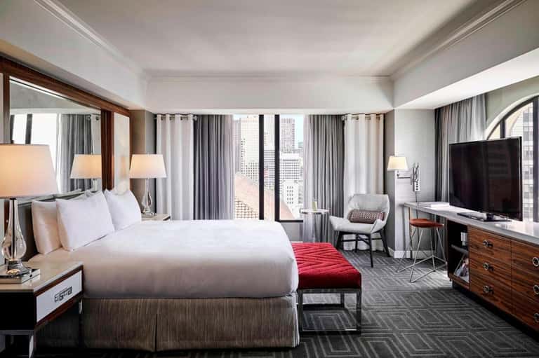 Luxury hotel bedroom with king bed, floor-to-ceiling windows, city skyline view, and modern furnishings