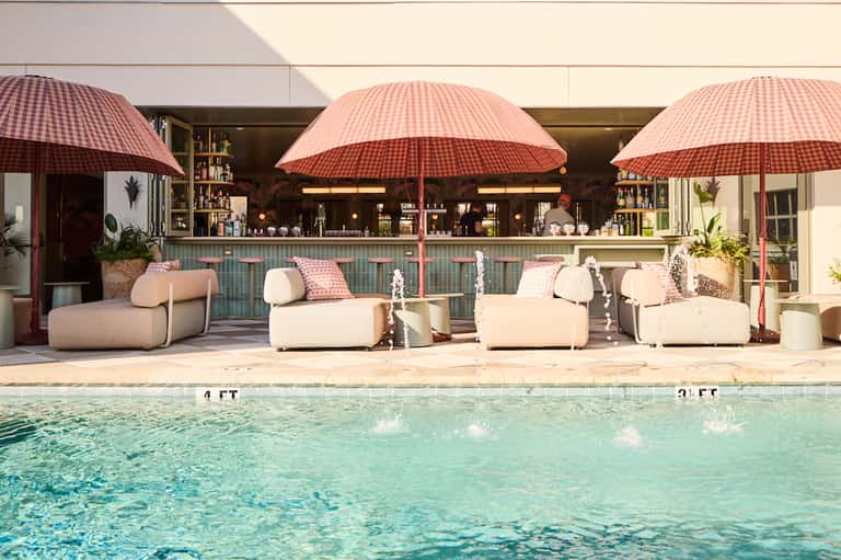 Resort poolside lounge with curved cream seating, red umbrellas, tiled bar, and crystal-clear water