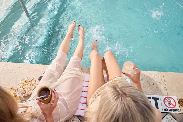 Person relaxing poolside with feet dangling over turquoise water, holding drink and snacks, no diving sign visible