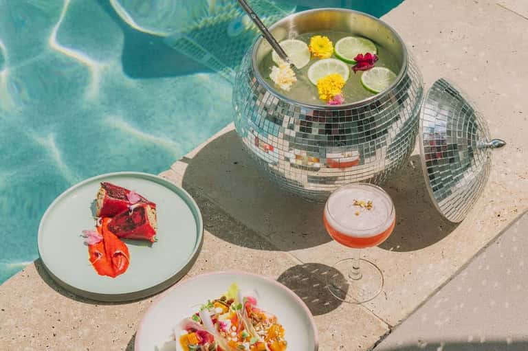 Pool-side gourmet dishes on white plates next to a decorative punch bowl with floating flowers