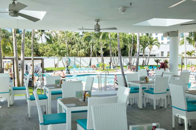 Modern covered dining area overlooking resort pool with white wicker furniture and tropical palm trees