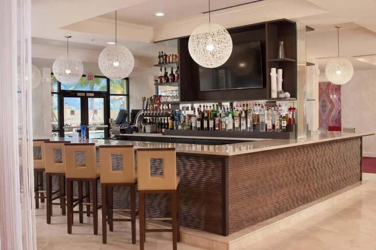Modern hotel bar with white countertop, dark shelving, woven pendant lights, and wooden bar stools with yellow cushions