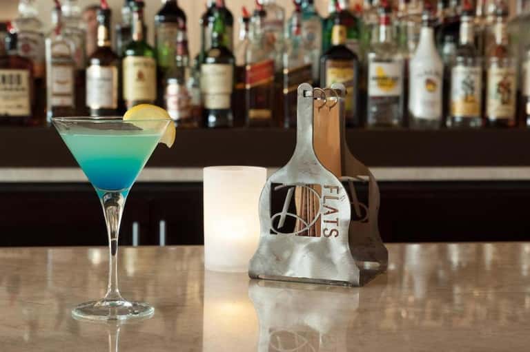 Turquoise cocktail with lemon slice on bar counter with full liquor bottle collection blurred in background