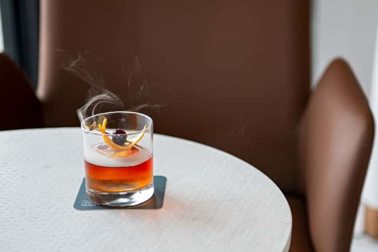 Cocktail with orange peel garnish and ice on a round table with brown background